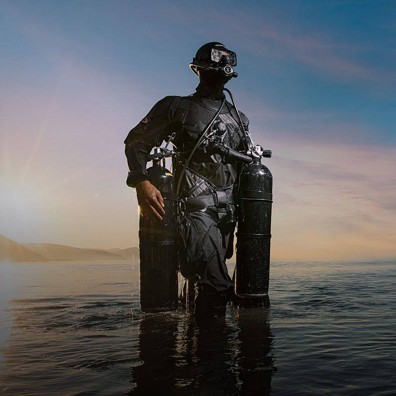 Diver wading into shallow water in a full drysuit with twin steel cylinders, sunset backdrop