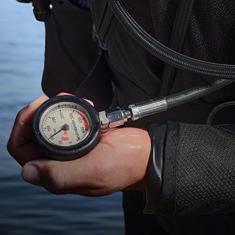 Diver's hand holding an analog pressure gauge attached to a coiled HP hose